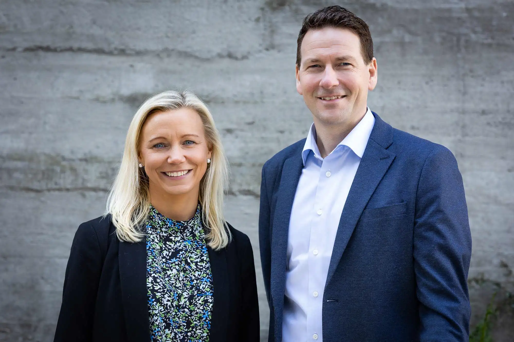 En kvinne og en mann i forretningsantrekk står ved siden av hverandre foran en betongvegg og smiler mot kamera