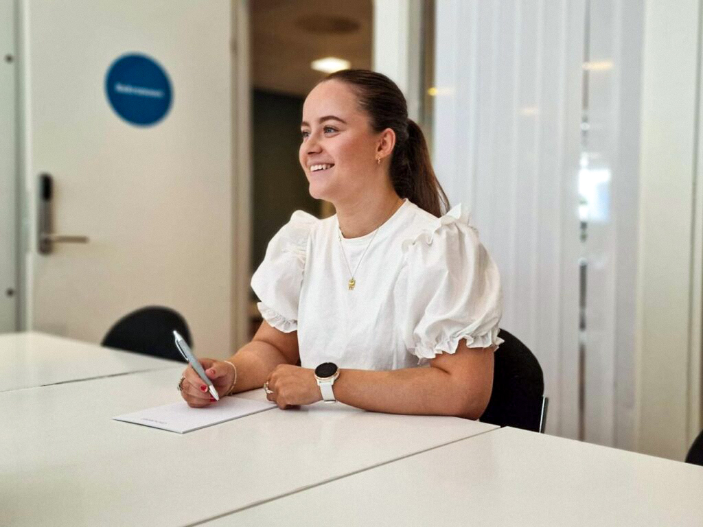 En kvinne i hvit bluse sitter ved et bord og skriver med en penn, smiler og ser til siden